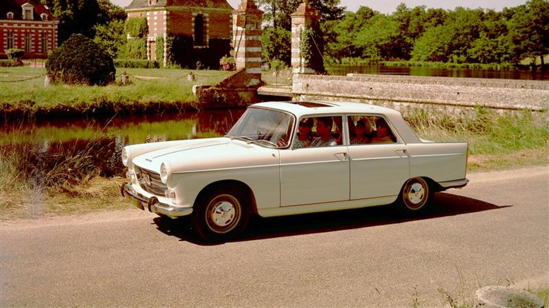 PEUGEOT 404 sur une route de campagne devant un château en brique rouge et verdure